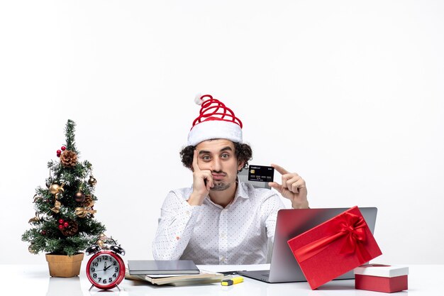 Estado de ánimo de Navidad con joven barbudo empresario pensativo con sombrero de santa claus con tarjeta bancaria sobre fondo blanco.