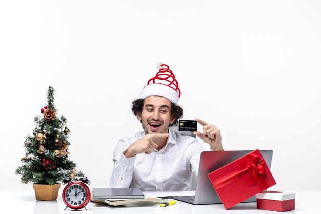 Estado de ánimo de Navidad con joven empresario positivo barbudo con sombrero de santa claus sosteniendo y señalando la tarjeta bancaria sobre fondo blanco.