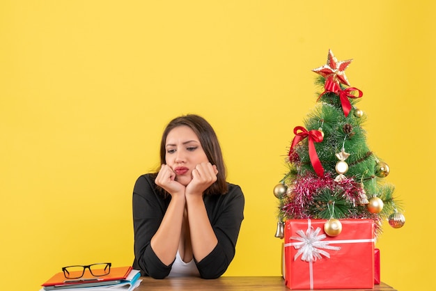 Estado de ánimo de Navidad con joven hermosa mujer seria centrada en algo cuidadosamente sentado en la oficina en amarillo