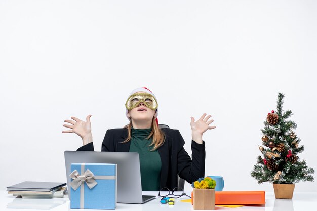 Estado de ánimo de Navidad con una mujer joven con gorro de Papá Noel y máscara con máscara sentado en una mesa mirando hacia arriba sobre un fondo blanco.