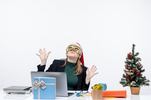 Estado de ánimo de Navidad con una mujer joven con gorro de Papá Noel y máscara con máscara sentado en una mesa mirando hacia arriba sobre un fondo blanco.