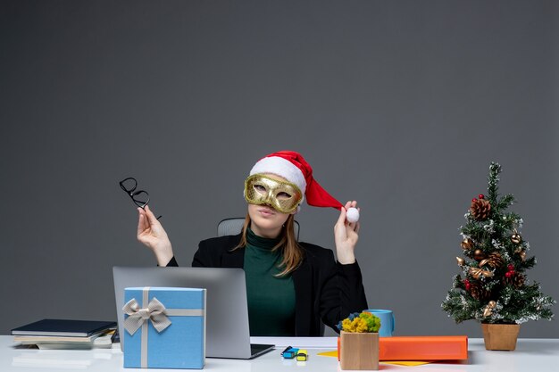 Estado de ánimo de Navidad con mujer joven jugando con sombrero de santa claus sosteniendo anteojos y vistiendo máscara sentado en una mesa sobre fondo oscuro