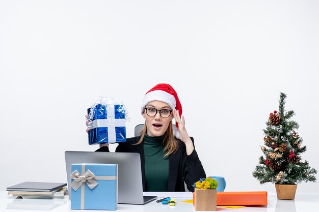Estado de ánimo de Navidad con mujer joven segura con sombrero de santa claus y gafas sentado en una mesa sosteniendo su regalo sobre fondo blanco.