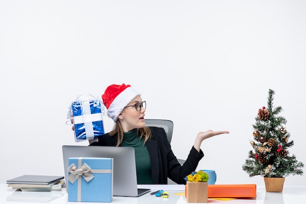 Estado de ánimo de Navidad con mujer joven con sombrero de santa claus y gafas sentado en una mesa sosteniendo su regalo haciendo preguntas sobre fondo blanco.