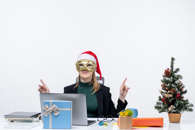 Estado de ánimo de Navidad con una mujer joven con sombrero de santa claus y con máscara sentado en una mesa apuntando algo sobre fondo blanco.