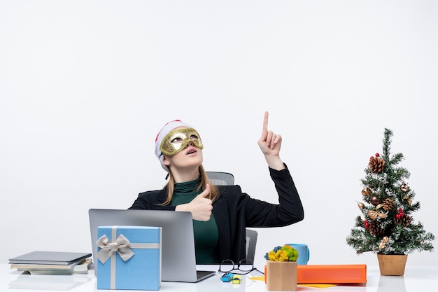 Foto gratuita estado de ánimo de navidad con una mujer joven con sombrero de santa claus y con máscara sentado en una mesa apuntando arriba sobre fondo blanco.