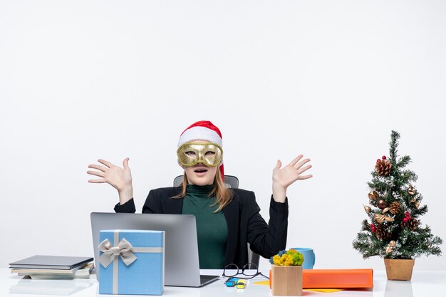 Estado de ánimo de Navidad con una mujer joven sorprendida con sombrero de santa claus y con máscara sentado en una mesa sobre un fondo blanco.