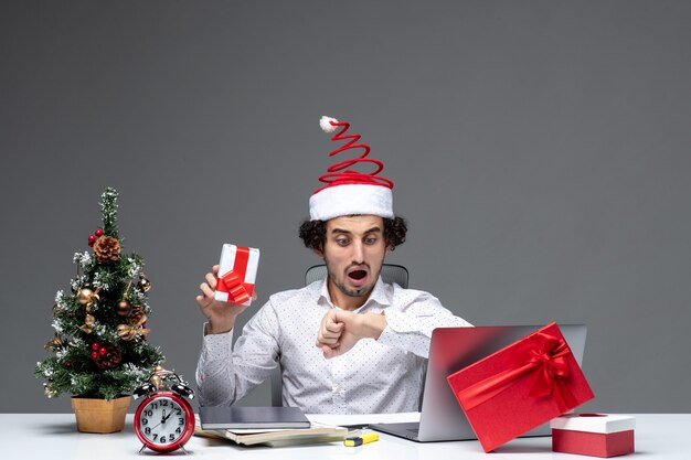 Estado de ánimo de Navidad con nervioso joven empresario con sombrero de santa claus sosteniendo su regalo y controlando su tiempo sobre fondo oscuro