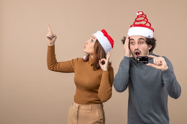 Estado de ánimo de Navidad con una pareja fresca confundida emocionada con sombreros rojos de santa claus chico mostrando tarjeta bancaria. mujer apuntando arriba