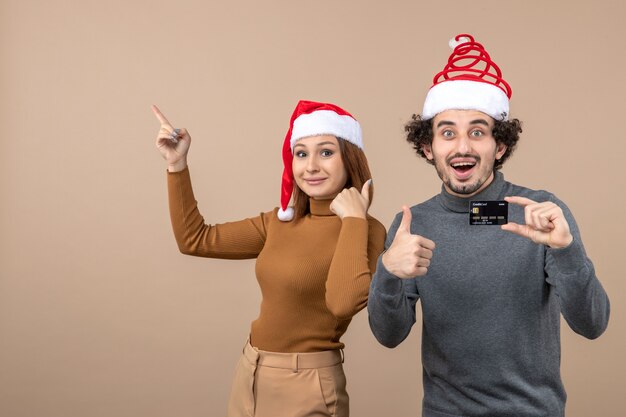 Estado de ánimo de Navidad con pareja fresca satisfecha emocionado vistiendo sombreros rojos de santa claus chico mostrando tarjeta bancaria, mujer apuntando por encima de ambos haciendo un gesto perfecto