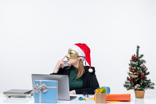 Estado de ánimo de Navidad con pensativo joven con sombrero de santa claus y máscara con máscara sentado en una mesa sobre fondo blanco.