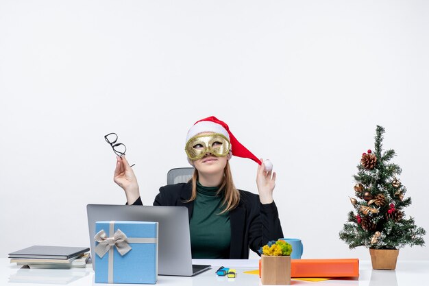 Estado de ánimo de Navidad con soñadora joven jugando con sombrero de santa claus sosteniendo anteojos y vistiendo máscara sentado en una mesa sobre fondo blanco.