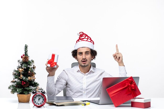 Estado de ánimo de Navidad con sonriente joven empresario con sombrero de santa claus y sosteniendo su regalo apuntando arriba sobre fondo blanco.