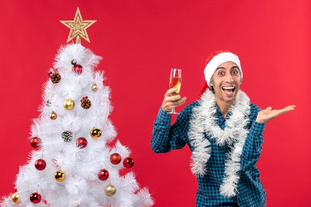 Estado de ánimo de Navidad con sonriente joven feliz con sombrero de santa claus en una camisa azul a rayas sosteniendo una copa de vino cerca del árbol de Navidad