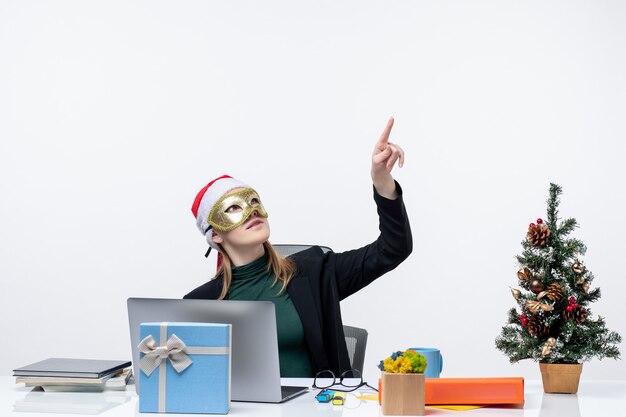 Estado de ánimo de Navidad con tensa joven con sombrero de santa claus y máscara con máscara sentado en una mesa apuntando hacia arriba sobre un fondo blanco.