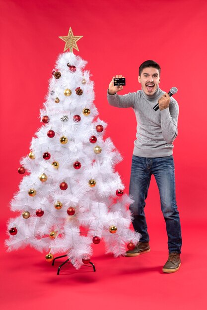 Estado de ánimo de Navidad con un tipo orgulloso de pie cerca del árbol de Navidad decorado y sosteniendo el micrófono y el teléfono