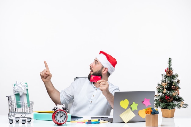 Estado de ánimo navideño con un adulto joven con sombrero de santa claus y sosteniendo una tarjeta bancaria y apuntando arriba a la izquierda sorprendentemente en la oficina