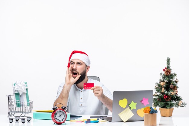 Estado de ánimo navideño con un adulto joven con sombrero de santa claus y sosteniendo una tarjeta bancaria y gritando a alguien en la oficina
