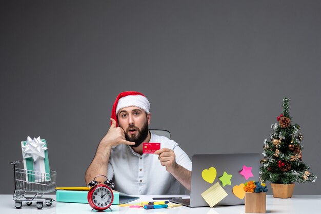 Estado de ánimo navideño con un adulto joven con sombrero de santa claus y sosteniendo una tarjeta bancaria sorprendentemente y haciendo un gesto de llamarme en la oficina