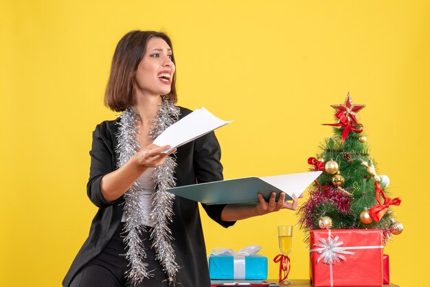 Estado de ánimo navideño con una bella dama sonriente de pie en la oficina e investigando documentos haciendo preguntas