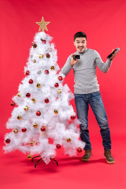 Estado de ánimo navideño con un chico emocional de pie cerca del árbol de Navidad decorado y sosteniendo el micrófono y el teléfono y hablando