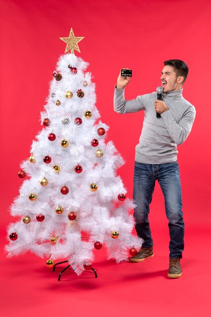 Estado de ánimo navideño con un chico seguro vestido con jeans de pie cerca del árbol de Navidad decorado y sosteniendo el micrófono grabando video