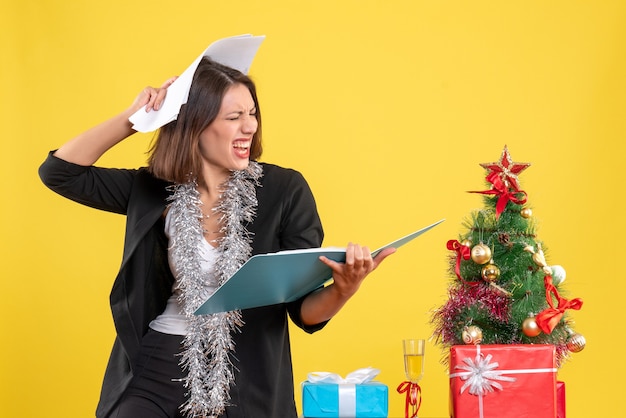 Estado de ánimo navideño con hermosa dama emocional de pie en la oficina e investigando documentos en amarillo