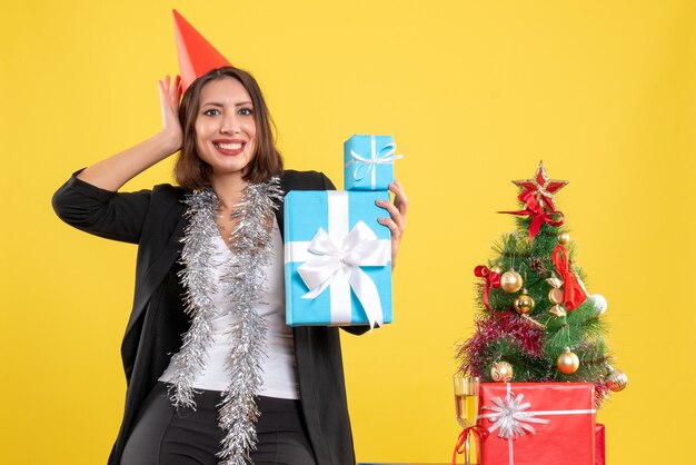 Estado de ánimo navideño con hermosa dama positiva con sombrero de Navidad con regalos en la oficina en amarillo
