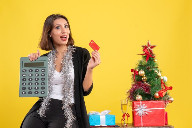 Estado de ánimo navideño con hermosa dama sonriente de pie en la oficina y sosteniendo la tarjeta bancaria calculadora en la oficina en amarillo