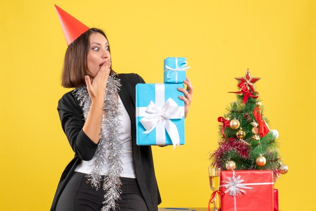 Estado de ánimo navideño con hermosa dama sorprendida con sombrero de Navidad con regalos en la oficina en amarillo