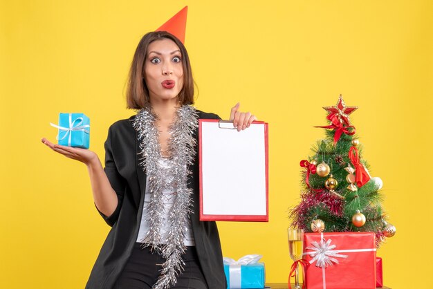 Estado de ánimo navideño con hermosa dama sorprendida sosteniendo documento y regalo en la oficina en amarillo