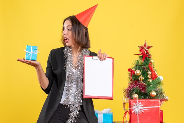 Estado de ánimo navideño con hermosa dama sorprendida sosteniendo documento y regalo en la oficina en amarillo
