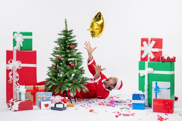 Estado de ánimo navideño con joven santa claus acostado detrás del árbol de navidad cerca de regalos en diferentes colores sobre fondo blanco imagen