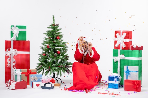 Estado de ánimo navideño con joven santa claus asustado sentado cerca del árbol de navidad y regalos en diferentes colores sobre fondo blanco.