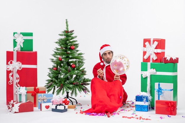 Estado de ánimo navideño con joven santa claus sentado cerca del árbol de navidad y regalos en diferentes colores sobre fondo blanco.