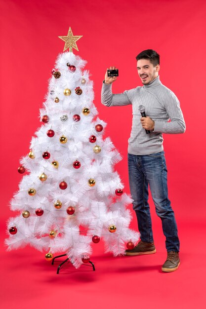 Estado de ánimo navideño con un joven sonriente de pie cerca del árbol de Navidad decorado y sosteniendo el micrófono y el teléfono