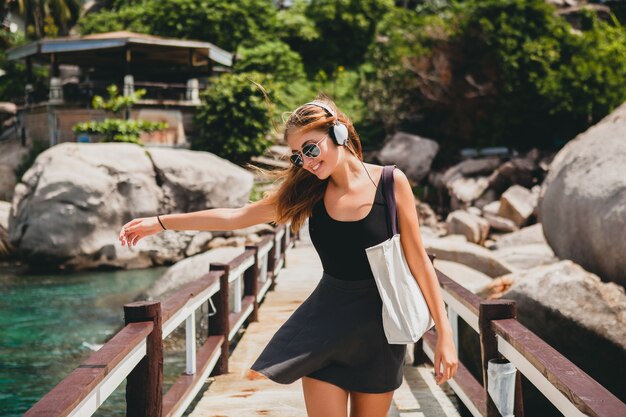 Estado de ánimo positivo de la mujer joven con estilo hipster, sonriendo feliz, viajando por el mundo, gafas de sol, auriculares, escuchando música, vacaciones de verano tropical, falda, disfrutar de la libertad, coqueta, sexy