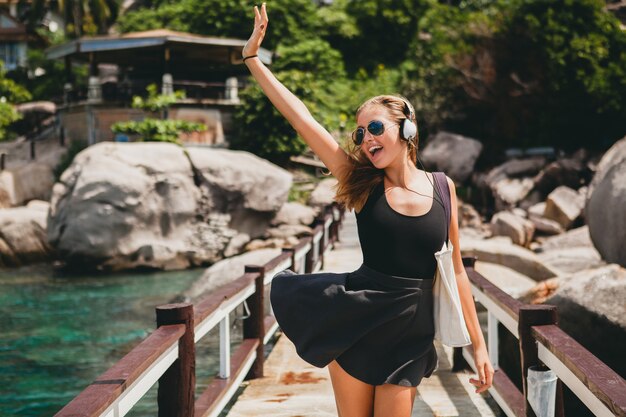 Estado de ánimo positivo de la mujer joven con estilo hipster, sonriendo feliz, viajando por el mundo, gafas de sol, auriculares, escuchando música, vacaciones de verano tropical, falda, disfrutar de la libertad, coqueta, sexy