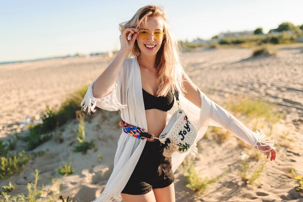 Estado de ánimo de verano. Elegante y despreocupada chica rubia en la playa de moda boho con flecos y accesorios divirtiéndose en la increíble playa solitaria.