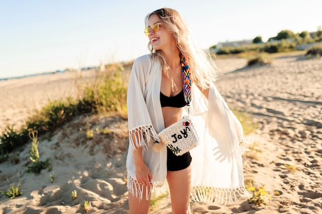 Foto gratuita estado de ánimo de verano. elegante y despreocupada chica rubia en la playa de moda boho con flecos y accesorios divirtiéndose en la increíble playa solitaria.