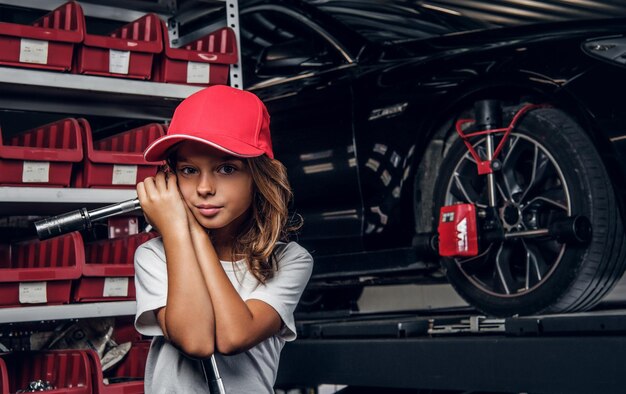 Estante con herramientas, auto brillante y niña con sombrero con una gran llave en las manos.