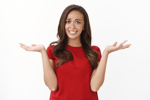 Estás bromeando Frustrado y molesto enloquecido mujer morena molesta con camiseta roja encogiéndose de hombros levanta las manos decepcionado encogerse escuchar tonterías estúpidas extrañas acusaciones fondo blanco