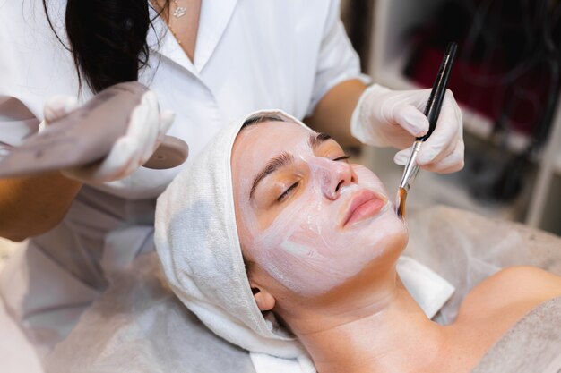 Esteticista con un pincel aplica una mascarilla hidratante blanca a la cara de un cliente joven en un salón de belleza spa