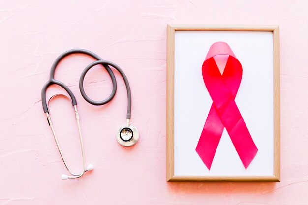 Estetoscopio cerca del marco de madera con cinta rosa de la conciencia del cáncer de mama