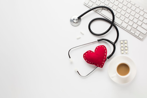 Estetoscopio con corazón rojo cerca de medicamentos; taza de café y teclado sobre escritorio blanco