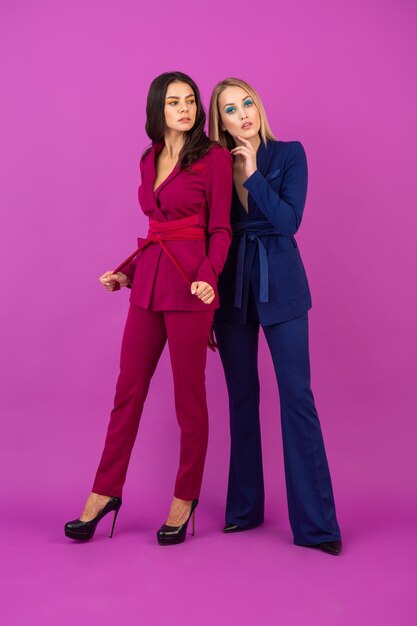 Estilo de alta moda dos mujeres atractivas en la pared violeta en elegantes y coloridos trajes de noche de color púrpura y azul, amigos divirtiéndose juntos, tendencia de la moda