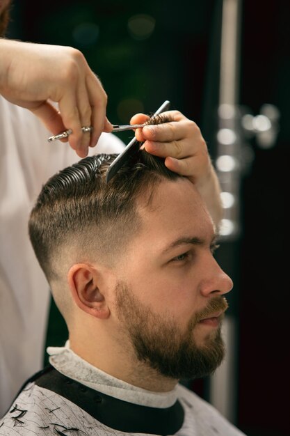 Estilo fresco. Primer plano del cliente del maestro barbero, estilista durante la atención y el nuevo aspecto del peinado. Ocupación profesional, belleza masculina y concepto de autocuidado. Colores suaves y enfoque, vintage.