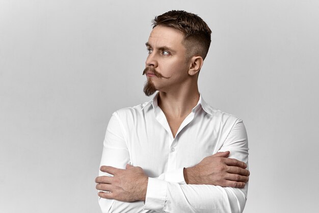 Estilo, moda y concepto de negocio. Imagen de estudio de confianza elegante joven empresario europeo con elegante bigote y barba cruzando los brazos sobre el pecho y mirando a otro lado, con mirada pensativa