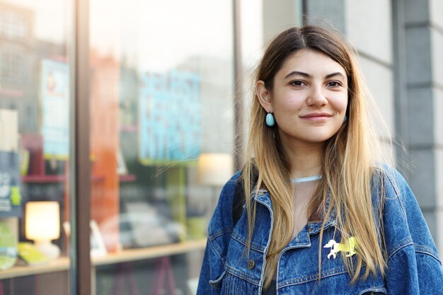 Estilo y moda. Tiro urbano de hermosa joven encantadora con cabello rubio vestida con una chaqueta de mezclilla de moda mientras compra en el centro de la ciudad, mirando y sonriendo, disfrutando del tiempo libre