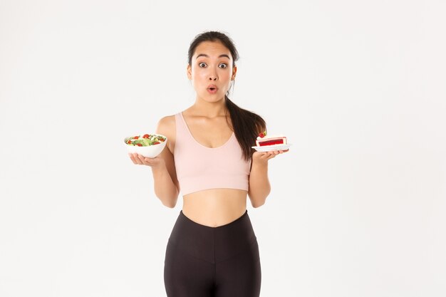 Estilo de vida activo, fitness y concepto de bienestar. Retrato de linda chica asiática indecisa y tentadora tratando de resistir la tentación mientras sostiene un delicioso pastel, está a dieta, mirando una ensalada saludable.